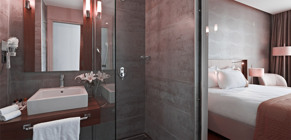 A hotel room with a bathroom and shower, a bed with white linens and a pink blanket, and a glass door separating the bathroom from the rest of the room.